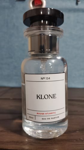 Reviewer's clear glass bottle of Klone Rouge Atlantic perfume, showing the white label with a black cap on a wooden surface.