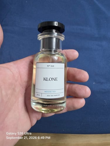 Customer's hand holding a clear, glass Imagine This KLONE eau de parfum bottle against a dark blue background.