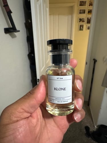 Customer's hand holding a clear, glass Imagine This Klone perfume bottle with a black cap.